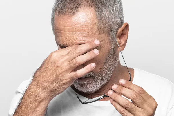 Older man experiencing eye irritation while holding glasses and rubbing his eyes due to discomfort.