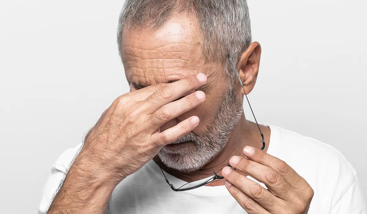 Older man experiencing eye irritation while holding glasses and rubbing his eyes due to discomfort.