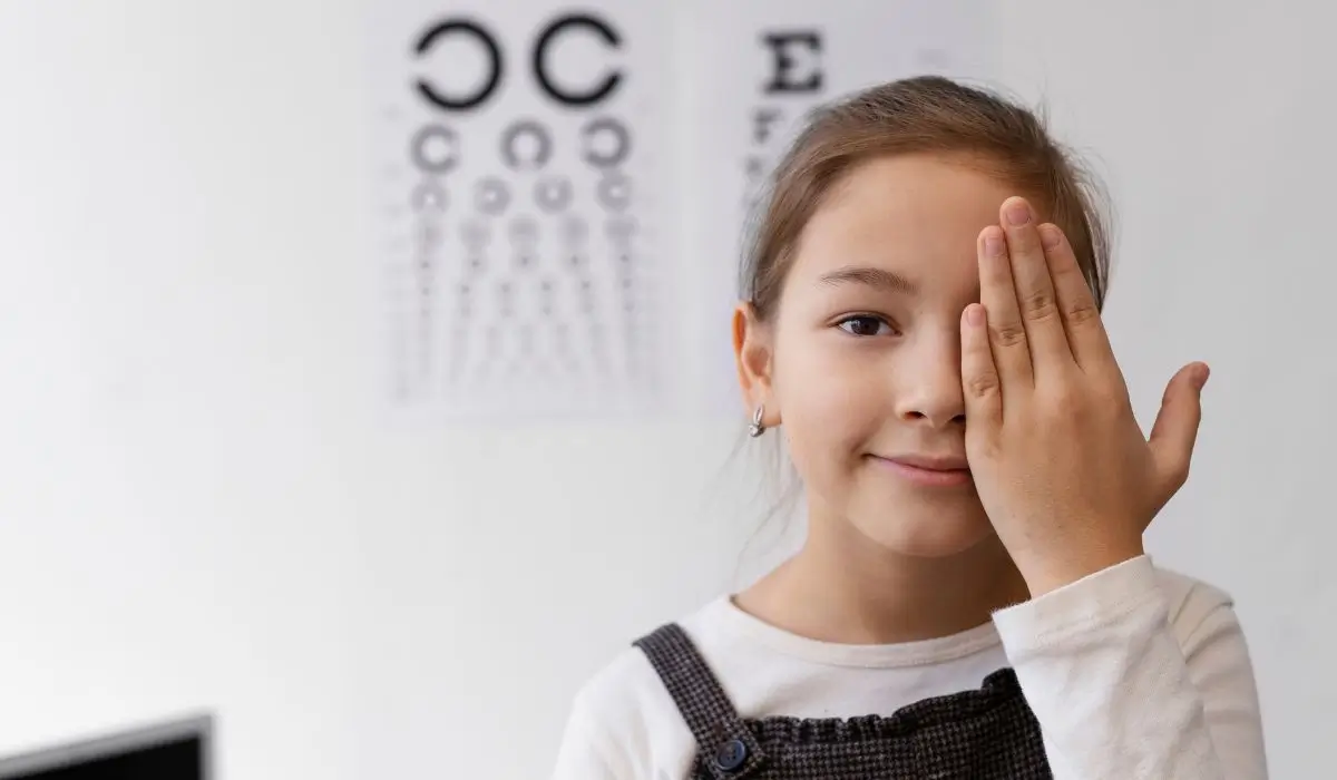 Child undergoing vision test to ensure pediatric eye health with eye chart in the background at clinic.