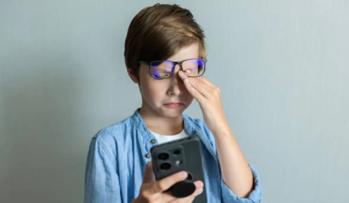 Young boy rubs his eyes while using a smartphone, showing effects of screen time and eye health concerns.